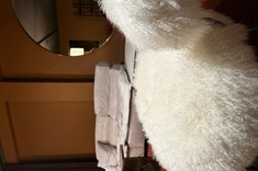 Kadr na fotel z białego futra, zapraszający do odpoczynku. Ciepłe tło i masywna, jasna sofa w głębi nadają wnętrzu przytulny i luksusowy charakter